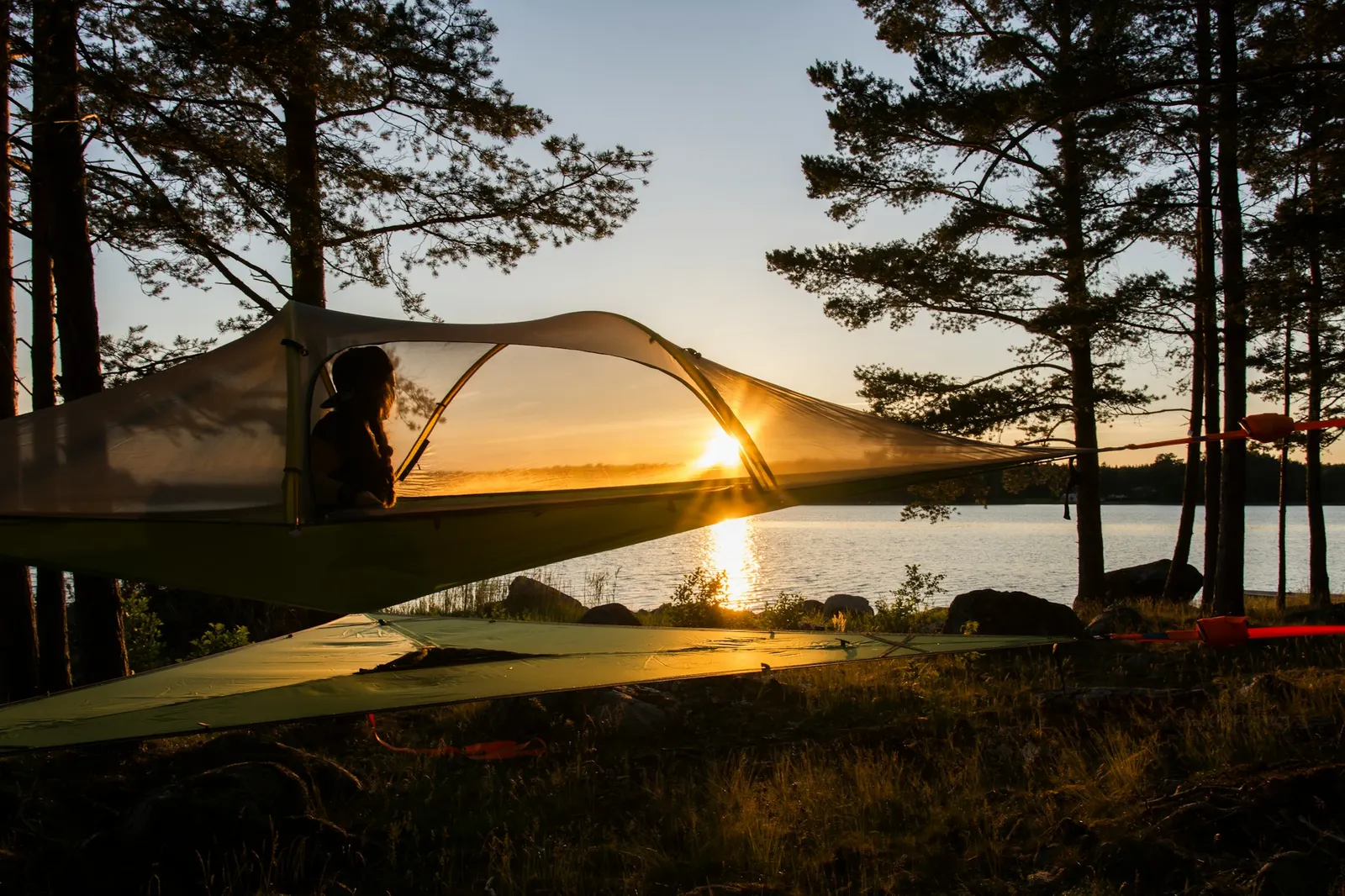 Suspended tree tent between pines at dusk with the sun setting over a lake