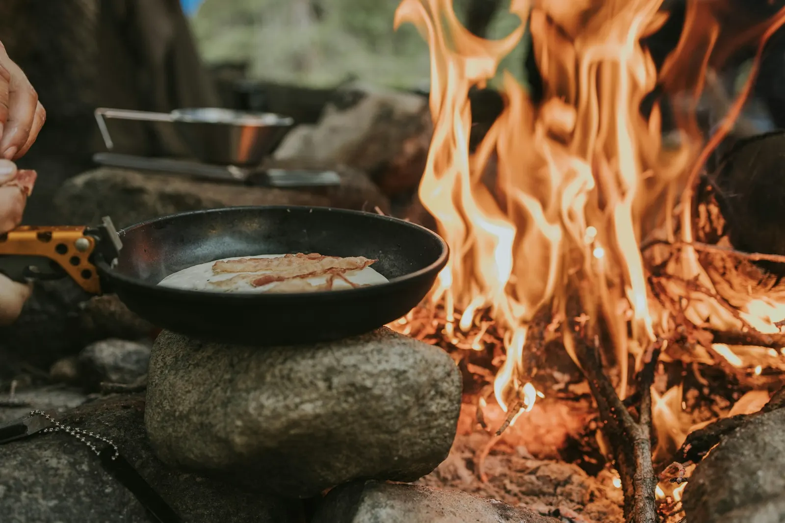 Cast-iron skillet with food cooking on rocks beside a lively campfire