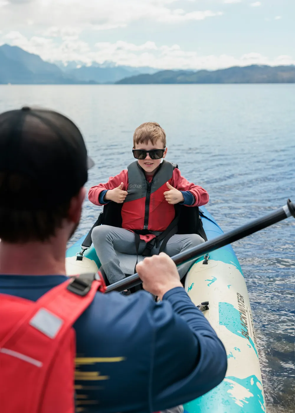 Smiling child in a red lifejacket giving thumbs up from a boat as a parent paddles