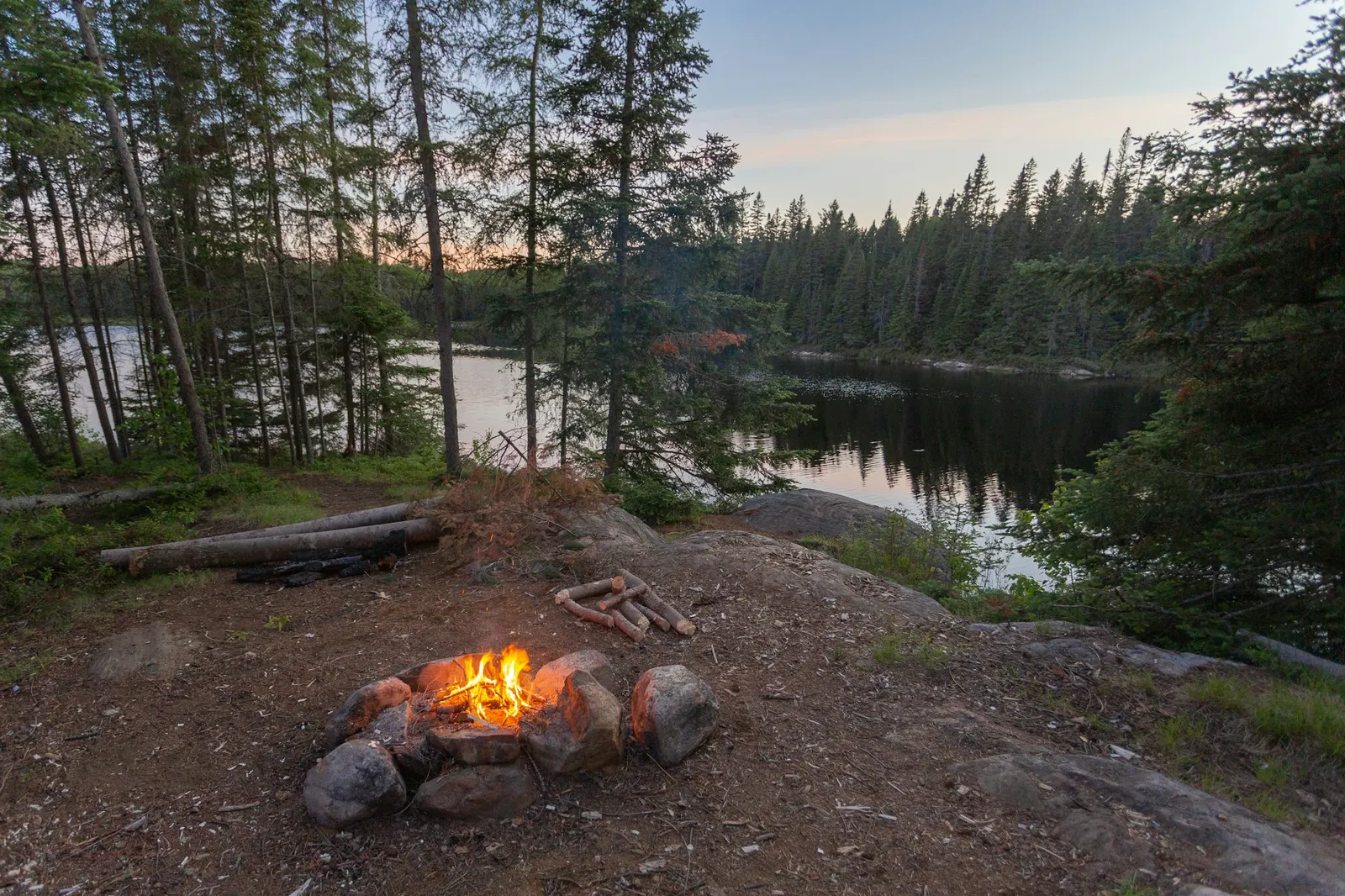 Small campfire in a stone ring on a rocky lakeshore campsite surrounded by pines