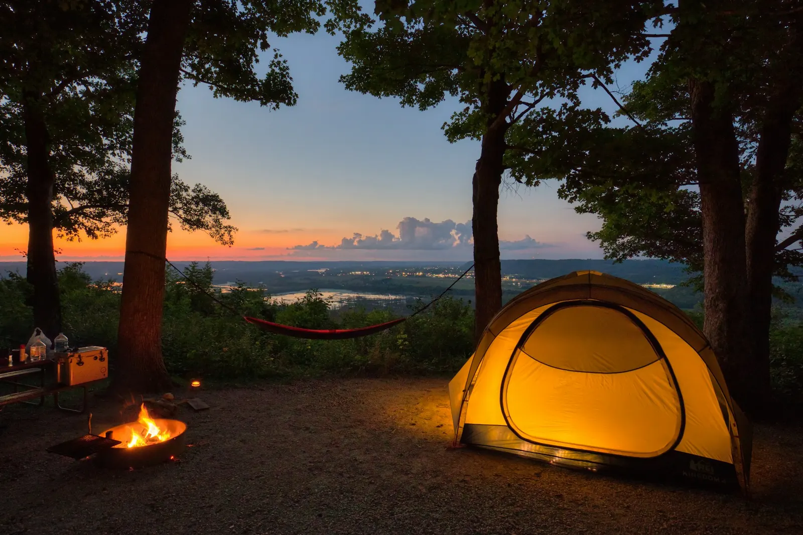 Yellow dome tent glowing at dusk beside a small campfire in a pine forest