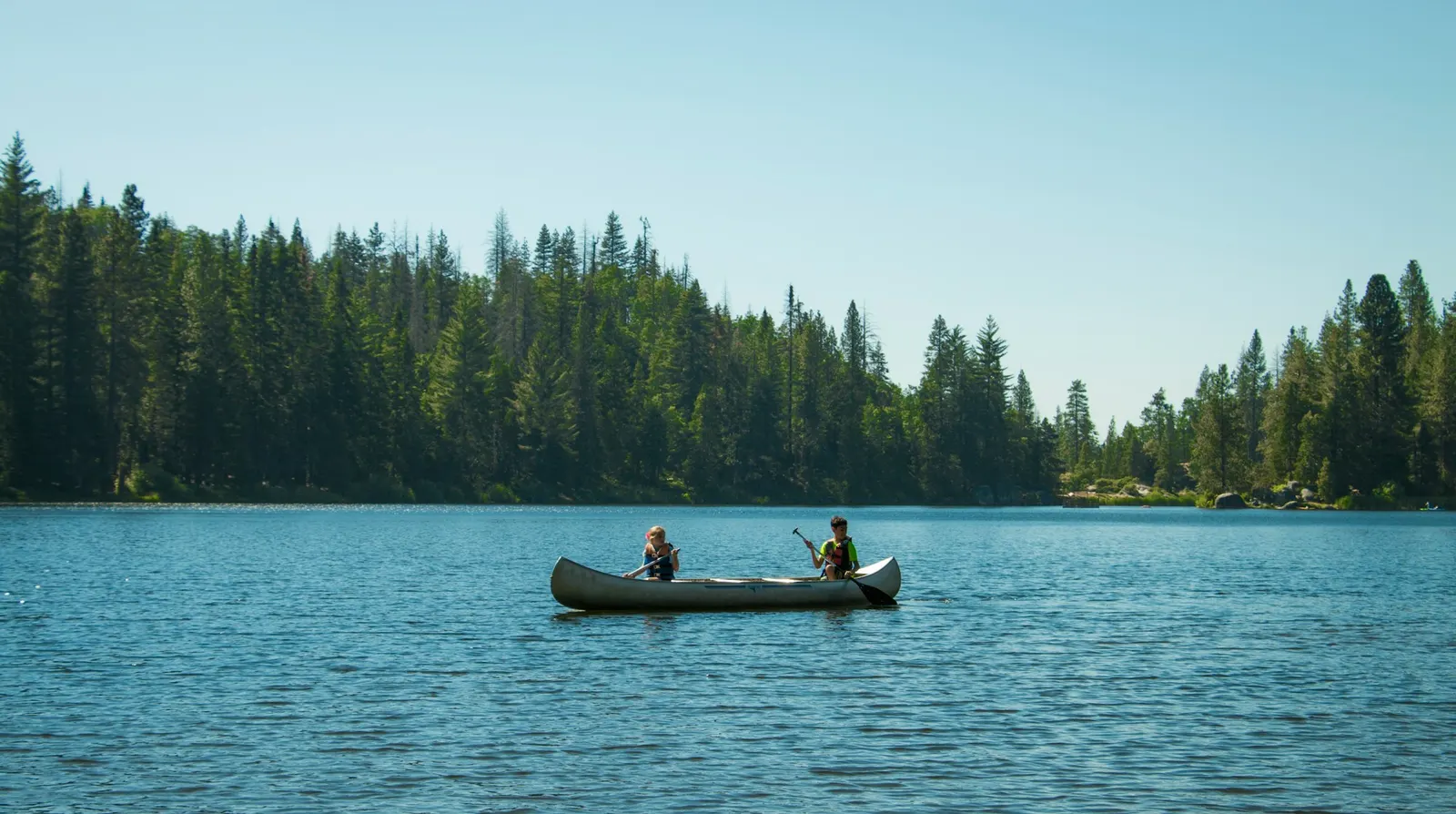 Two paddlers in a canoe crossing a calm lake lined with spruce forest