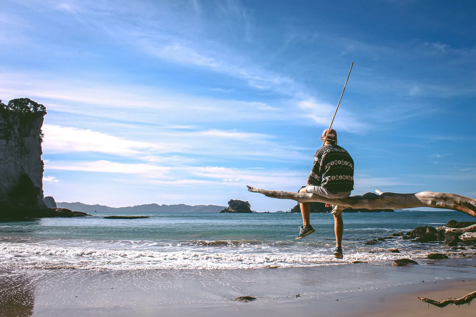 Person sitting on a large driftwood log on a sandy ocean beach under a blue sky